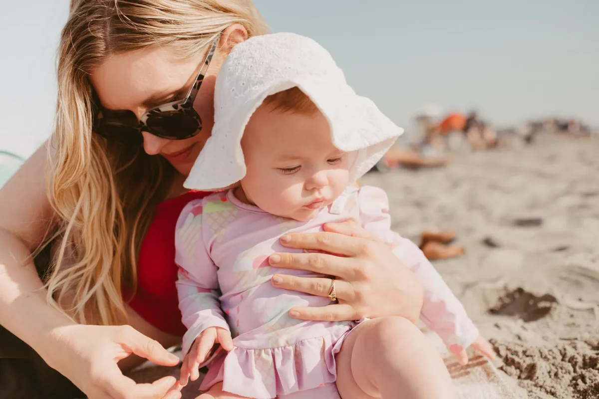 Mother applies sun protection for babies at the beach with a sun hat and long-sleeved clothing.