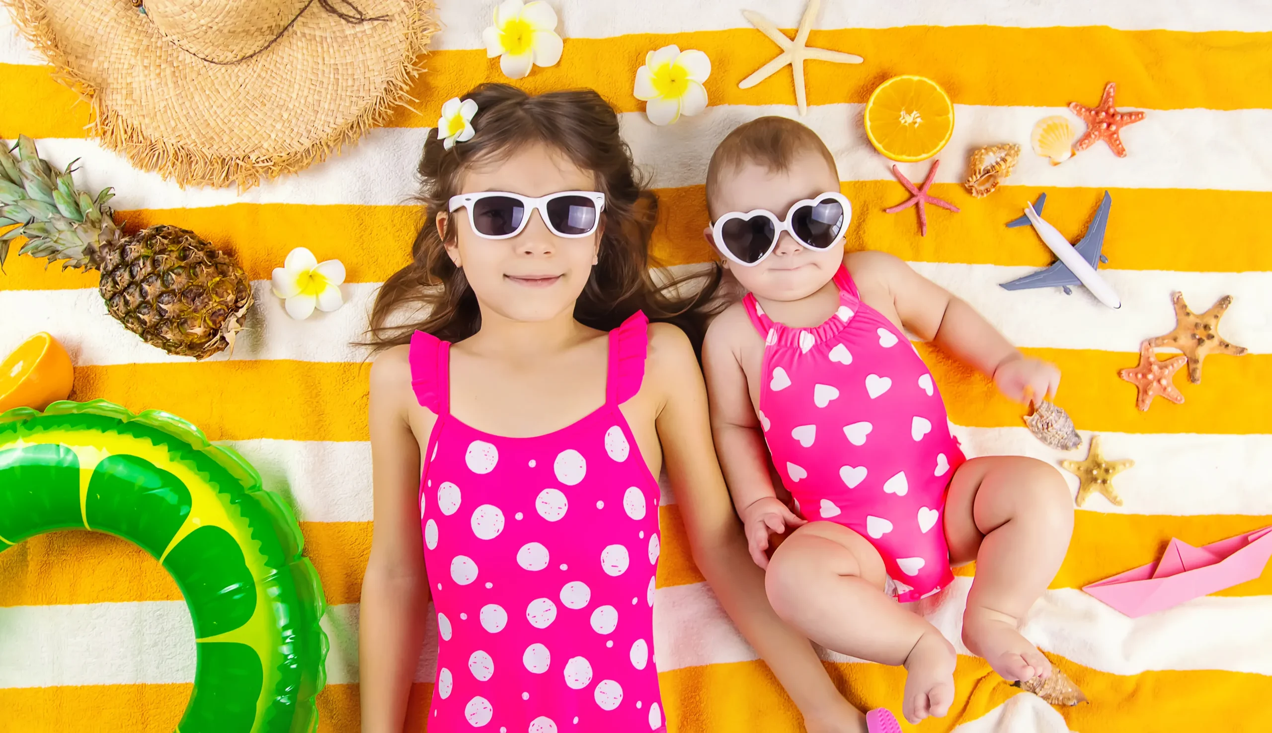 Two children in pink swimsuits and sunglasses lying on a striped towel—sun protection for babies with accessories and summer essentials.