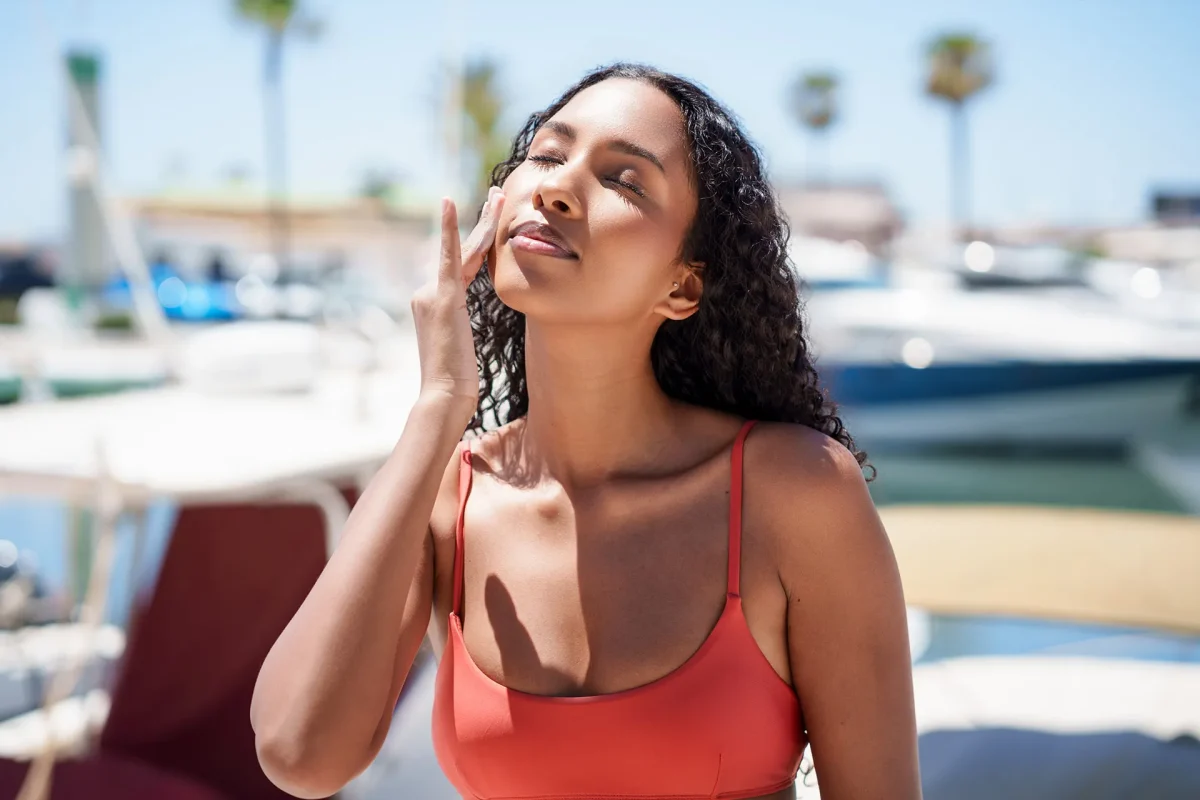 Woman with dark skin applying sunscreen to her face outdoors, demonstrating the importance of sunscreen for dark skin.