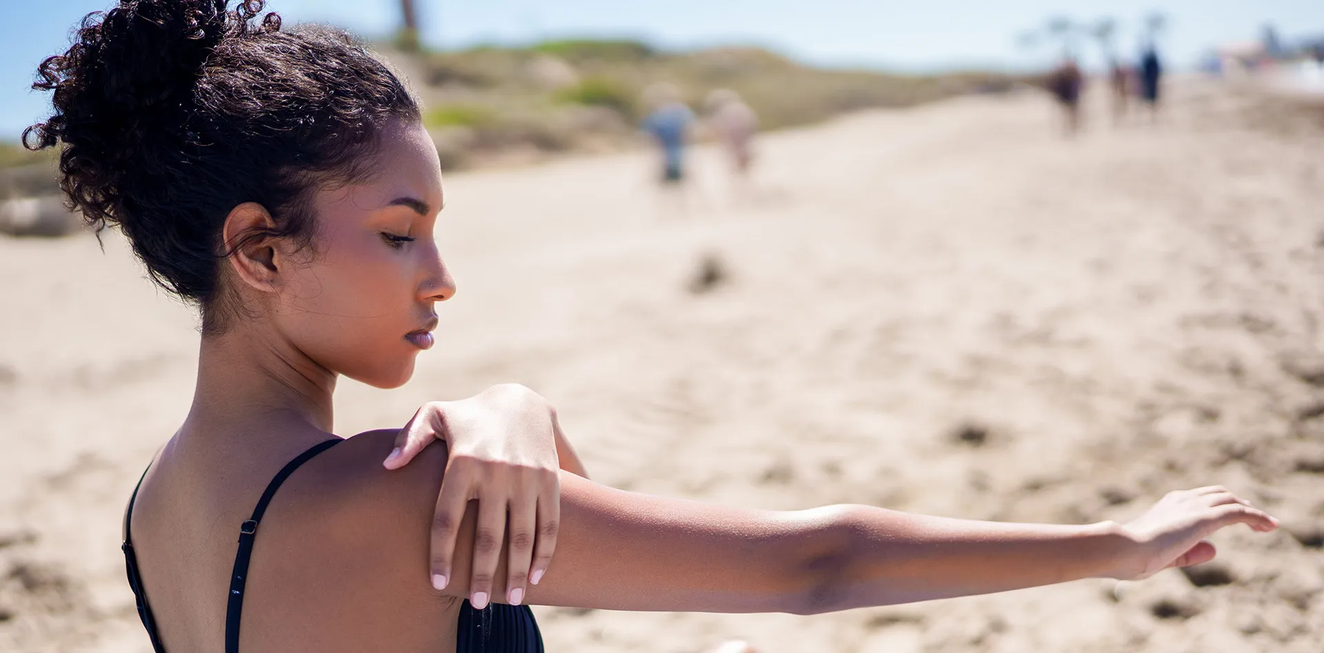 Young woman with dark skin applying sunscreen to her arm on a sunny beach, showing the importance of sunscreen for dark skin.