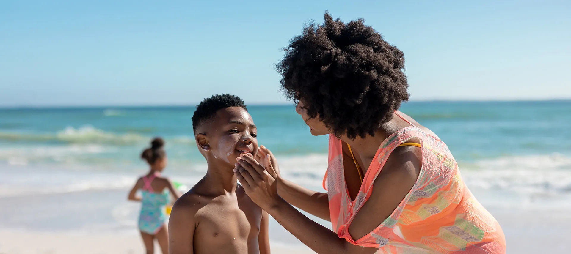 Mother applying sunscreen for dark skin to her son’s face on the beach, emphasizing the importance of sun protection for dark skin.
